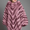 Model wearing a luxurious designer pink chinchilla poncho with diagonal chevron patterns, smiling confidently on a city sidewalk.