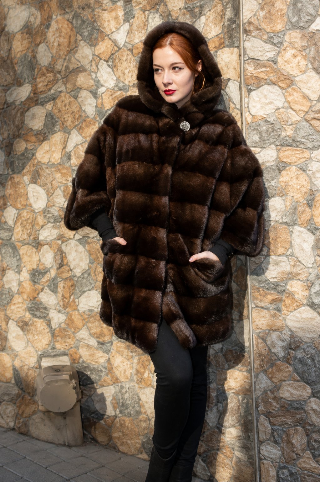 Demi mink jacket with hooded collar, cropped sleeves, and button closure, worn against a stone wall.