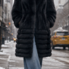 A stylish woman walking on a snowy New York City street at West 57th Street, wearing a plush black fox fur stroller coat with a tiered design, paired casually with wide-leg blue jeans, black pointed-toe boots, and dark sunglasses.
