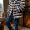 A smiling man standing by a lit stone fireplace wearing a luxurious grey and black horizontal-striped Chinchilla Rex Rabbit fur jacket with a large fluffy Silver Fox trim hood, currently on clearance.