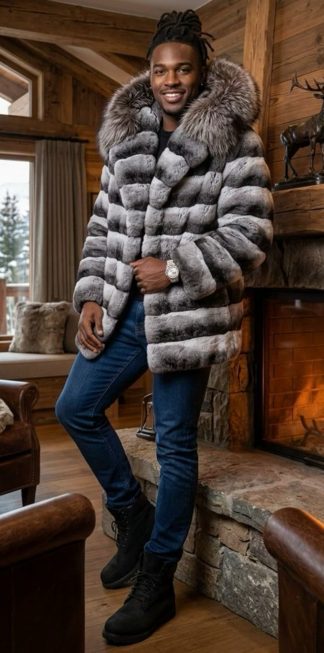 A smiling man standing by a lit stone fireplace wearing a luxurious grey and black horizontal-striped Chinchilla Rex Rabbit fur jacket with a large fluffy Silver Fox trim hood, currently on clearance.